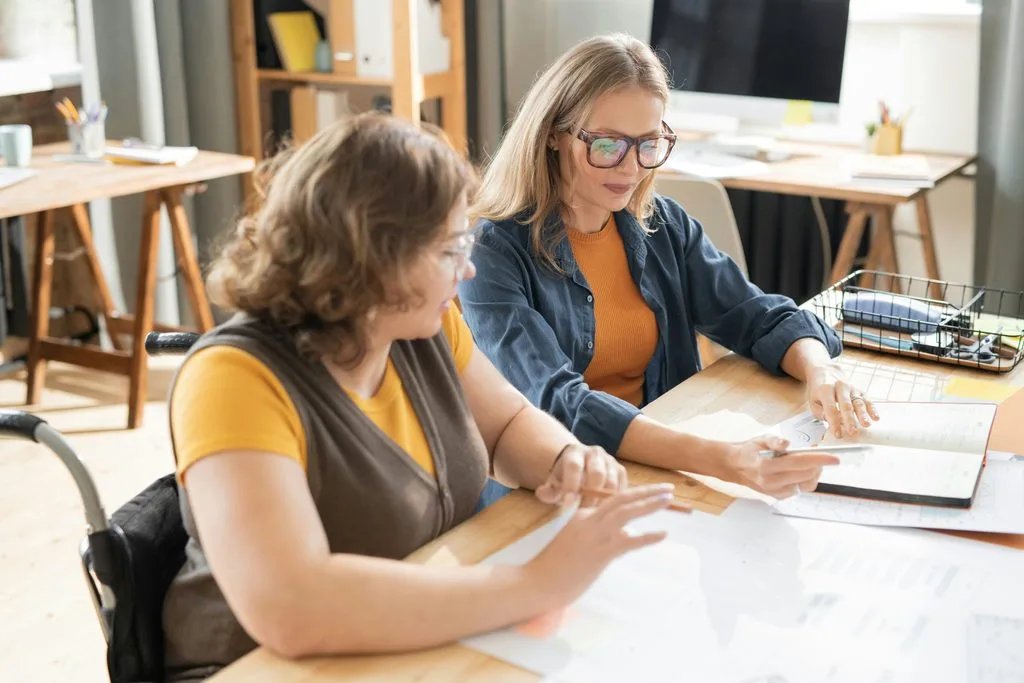 Two women review a planner and notes together at a table in a bright workspace; one uses a wheelchair.”