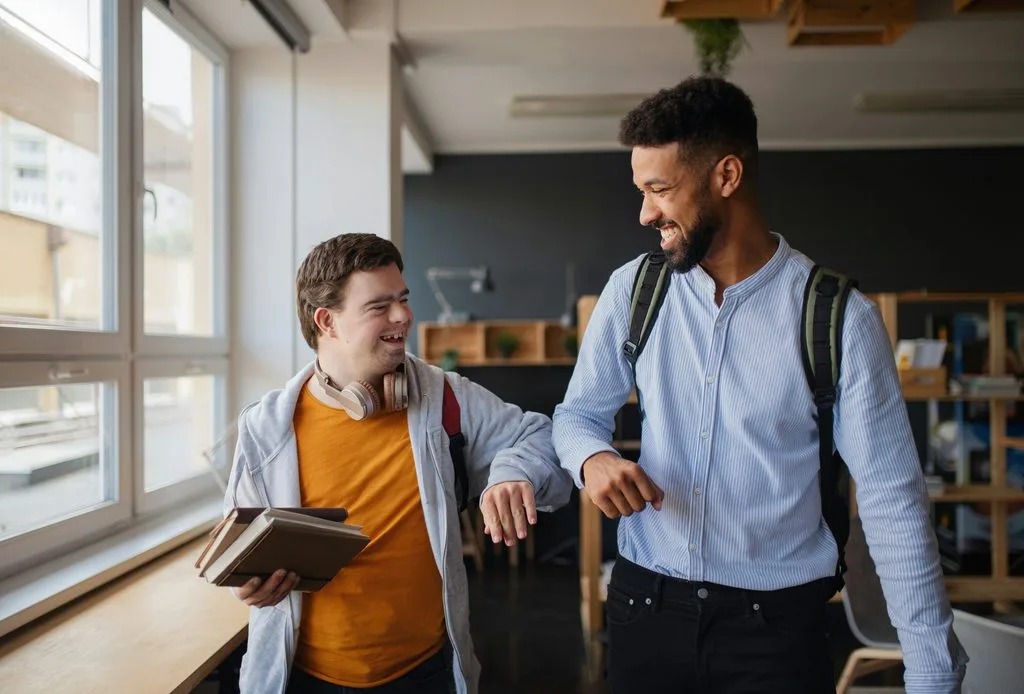 A young man holding books laughs with a friend carrying a backpack in a bright, sunlit room.